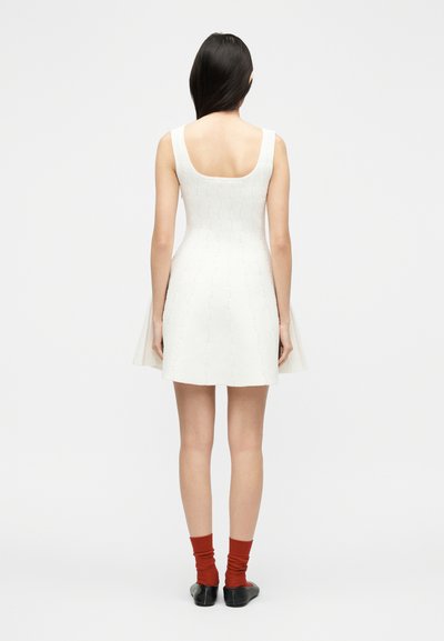 Γυναίκα που στέκεται με την πλάτη προς τον θεατή, φοράει ένα λευκό πλεκτό φόρεμα χωρίς μανίκια, κόκκινες κάλτσες και μαύρα μπαλαρίνες, μπροστά σε απλό λευκό φόντο.