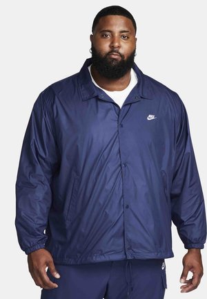 Giacca a vento Nike blu navy con collo a camicia, bottoni a pressione, tasche laterali e polsini elasticizzati. Logo visibile sul torace.