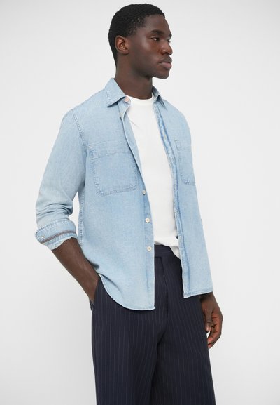 Hellblaues Jeanshemd mit Knopfleiste, zwei Brusttaschen und hochgekrempelten Ärmeln, kombiniert mit dunkler Hose mit Nadelstreifen.