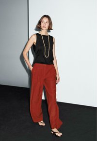 Black sleeveless top paired with loose red linen trousers and minimal black sandals, featuring a long bead necklace and a smooth texture.