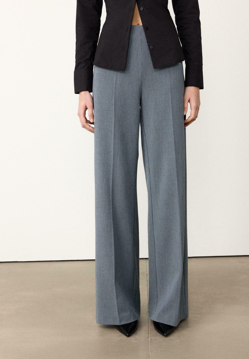 Pantalon large à fines rayures grises en tissu texturé, avec une taille haute, porté avec une chemise noire ajustée à boutons et des talons pointus.