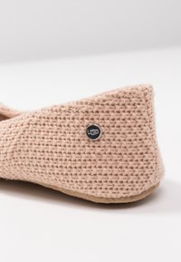 Detailní záběr na béžovou tkanou textilní papuču se semišovou podrážkou a malým kulatým kovovým logem UGG na patě.