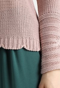Ljusrosa stickad tröja med texturerade ärmar och vågig fåll, i kombination med en mörkgrön kjol, som visar en mjuk, tjock känsla.