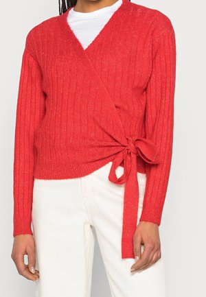 Red ribbed wrap sweater with a v-neck and tie detail at the waist, made of a textured knit fabric, paired with white pants.