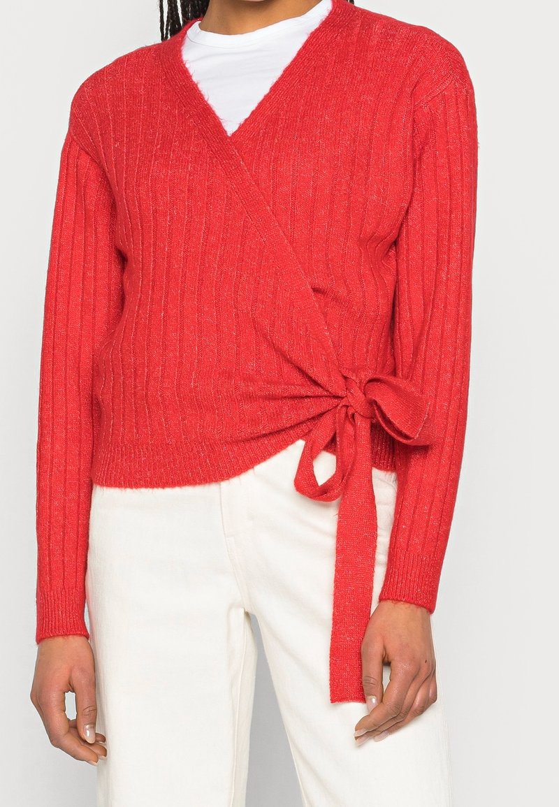 Suéter envolvente rojo de canalé con escote en V y detalle de lazo en la cintura, hecho de una tela de punto texturizada, combinado con pantalones blancos.