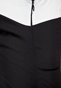 Svart och vit zip-up topp med dragkedja, som har en triangulär färgblockdesign och en mjuk, stretchig tygtextur.