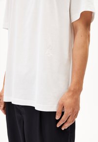 Witte katoenen t-shirt met korte mouwen, met een subtiele geborduurde detail nabij de zoom. Gestyleerd met losse, donkere broek.