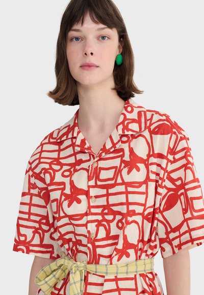 Femme aux cheveux bruns courts portant une chemise blanche avec des motifs graphiques rouges audacieux et une ceinture à carreaux jaunes nouée à la taille.