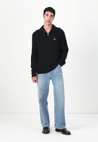 Maglione in maglia nero con zip e un piccolo logo sul petto a sinistra, abbinato a jeans dritti blu chiaro e scarpe nere.