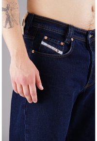 Dunkelblaue Jeans aus Denim verfügen über kupferne Nieten, ein Etikett an der Fronttasche und eine kontrastierende Naht. Der Stoff hat eine glatte Textur.