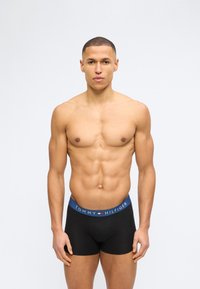 Μυώδης άνδρας με κοντά μαλλιά, που φοράει μαύρα μπαντόκια Tommy Hilfiger, στέκεται μπροστά σε ένα απλό φωτεινό φόντο.