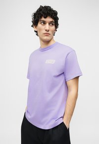 Νεαρός με σγουρά μαλλιά που φοράει λιλά κοντομάνικη μπλούζα με κείμενο στο στήθος και μαύρο παντελόνι, χέρια στις τσέπες, κοιτάζοντας πλάγια.