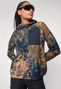 Fleece pullover in bruin en blauwe patronen, voorzien van een hoge kraag, een vo pocket en een gestructureerde stof. Model draagt donkere zonnebrillen.