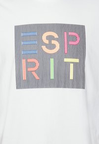 Camiseta blanca con un gráfico que presenta la palabra "ESPÍRITU" en letras coloridas y rayadas sobre un fondo de delgadas rayas verticales.