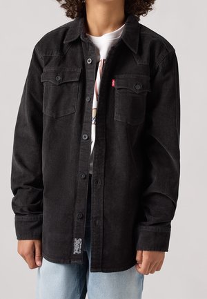 Osoba ubrana w czarną koszulę sztruksową na guziki, na białym t-shircie z nadrukiem i jasnoniebieskie dżinsy, z rękami swobodnie opuszczonymi wzdłuż ciała.