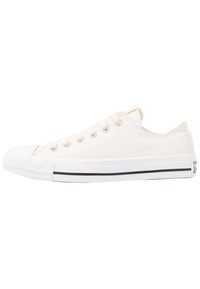 Baskets basses en toile couleur crème avec embout en caoutchouc, cinq œillets et bande noire sur la semelle extérieure. Tige en tissu texturé avec des accents contrastants.