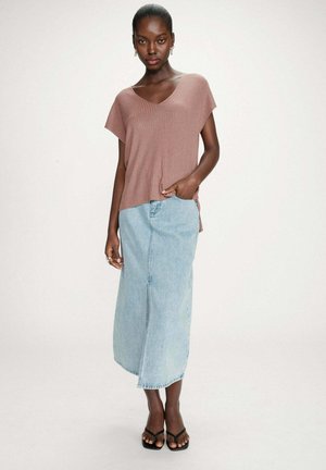 Femme portant un haut rose côtelé à manches courtes, rentré dans une jupe midi en denim bleu clair, avec des sandales noires à bouts carrés, debout devant un fond blanc.