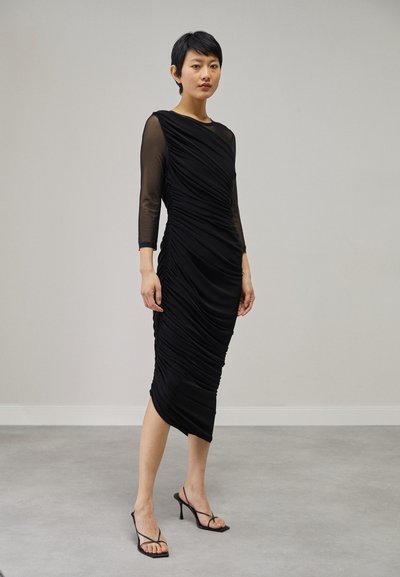 Schwarzes gerafftes Kleid mit transparenten langen Ärmeln, knielang, figurbetontes Design, glatte Textur, entspannter Ausschnitt, kombiniert mit schwarzen Riemchensandalen.