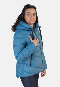 Chaqueta acolchada azul con capucha, con detalles de cremallera negra y diseño acolchado. Usada con jeans oscuros, mostrando vista frontal.