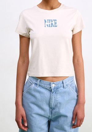 Mujer con camiseta corta Nike beige claro con logo azul y jeans de mezclilla azul claro de talle alto, de pie contra un fondo liso.