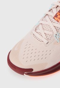 Baskets de sport rose clair avec une tige en mesh, fermeture à lacets et accents bordeaux. La semelle a une finition lisse avec des détails texturés.