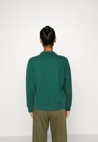 Felpa verde con colletto, spalle scese e polsini a coste. Indossata con pantaloni verde oliva, con una vestibilità rilassata e una consistenza morbida.