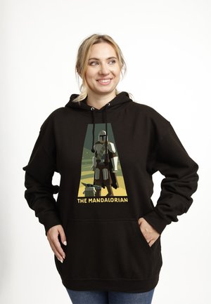 Femme souriante portant un sweat à capuche noir avec une illustration d’un Mandalorien blindé et d’un petit extraterrestre vert, intitulé « The Mandalorian ».