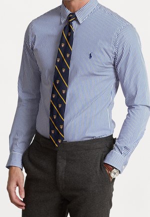 Mann, der ein blau-weiß gestreiftes Hemd, dunkelblaue Krawatte mit gelben Streifen und Wappen, graue Hose und eine Armbanduhr trägt.