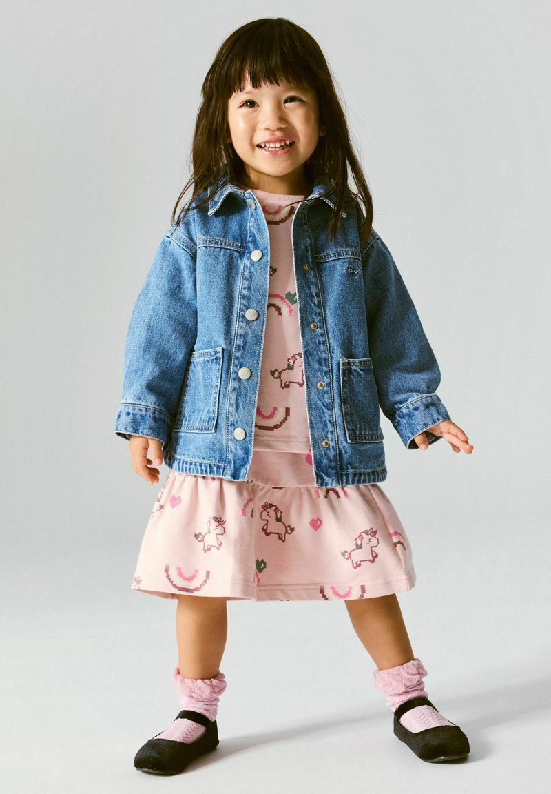 Jeune fille souriante portant une veste en jean bleue sur une robe rose avec des motifs de licornes et d'arc-en-ciel, des chaussettes roses et des chaussures noires, debout sur un fond clair.