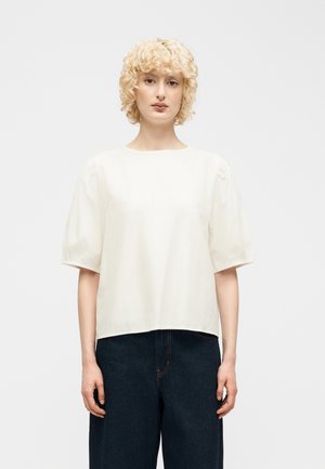 Personne aux cheveux blonds courts et bouclés, portant un chemisier blanc ample à manches bouffantes et un jean large taille haute de couleur foncée, se tenant droite.