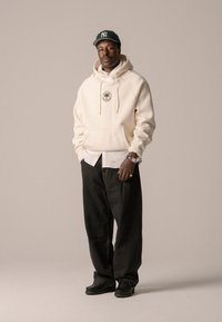 Cream hoodie with circular logo, layered over a white shirt. Black baggy jeans and black shoes complete the outfit, with a green cap.