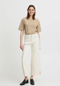 Beige bavlněné tričko a bílé široké džíny s bočními rozparky, doplněné černými mokasíny. Model stojí na jednobarevném pozadí.