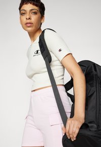 Svart ryggsekk med polstrede stropper. Lyst beige ribbet crop top med logo, kombinert med pastellrosa cargo-shorts. Glatte teksturer, uformell design.