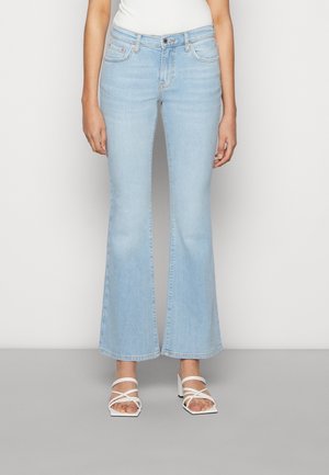 Woman wearing light blue high-waisted flare jeans and white strappy heeled sandals, standing against a plain light background.