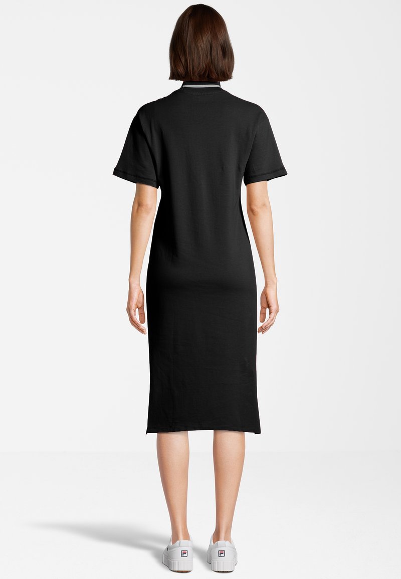 Donna in piedi di spalle, indossa un abito midi nero a maniche corte e sneakers bianche, su uno sfondo bianco semplice.