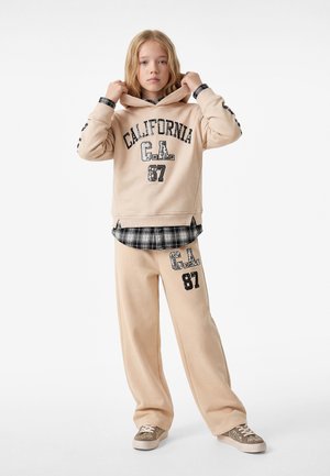 Ung person iført beige hættetrøje og bukser med teksten "California C.A. 87", lagdelt over en sort-hvid ternet skjorte, står stille.