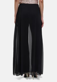 Jupe maxi en chiffon noir avec fentes sur les côtés, taille élastique, design fluide et texture superposée. Portée avec des chaussures à talons hauts.