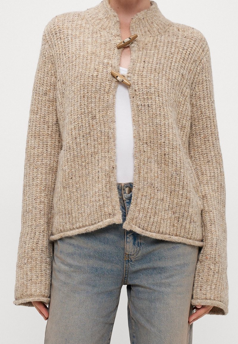 Beige stickad cardigan med ett texturerat mönster, med en ribbad krage och träspännen framtill. Löst passform, kort längd.