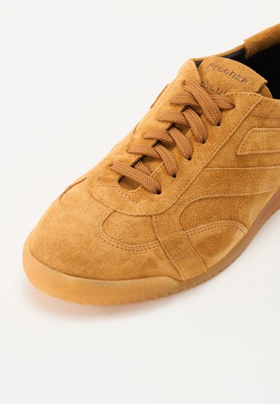 Zapatillas de ante color beige con una textura suave, puntera redonda, suela de goma y diseño de cordones texturizados. Presenta un logotipo en la lengüeta y acentos laterales.