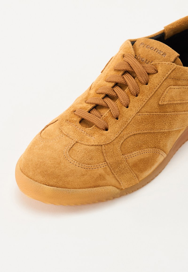 Tan suede sneaker with a smooth texture, round toe, rubber sole, and textured lace-up design. Features branding on the tongue and side accents.