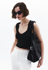 Top negro ajustado con diseño de escote en forma de V, combinado con pantalones blancos de cintura alta. La modelo sostiene un bolso negro con accesorios de acero inoxidable.