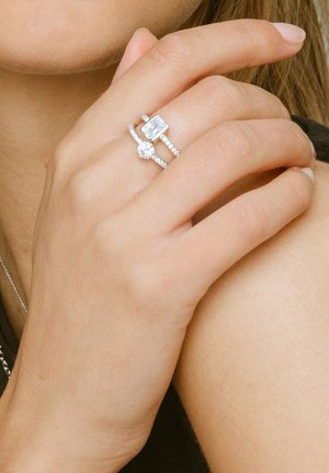 To sølvringer på en hånd: en rektangulær og en rund, begge sat med små klare sten, båret på en finger mod en bar arm.