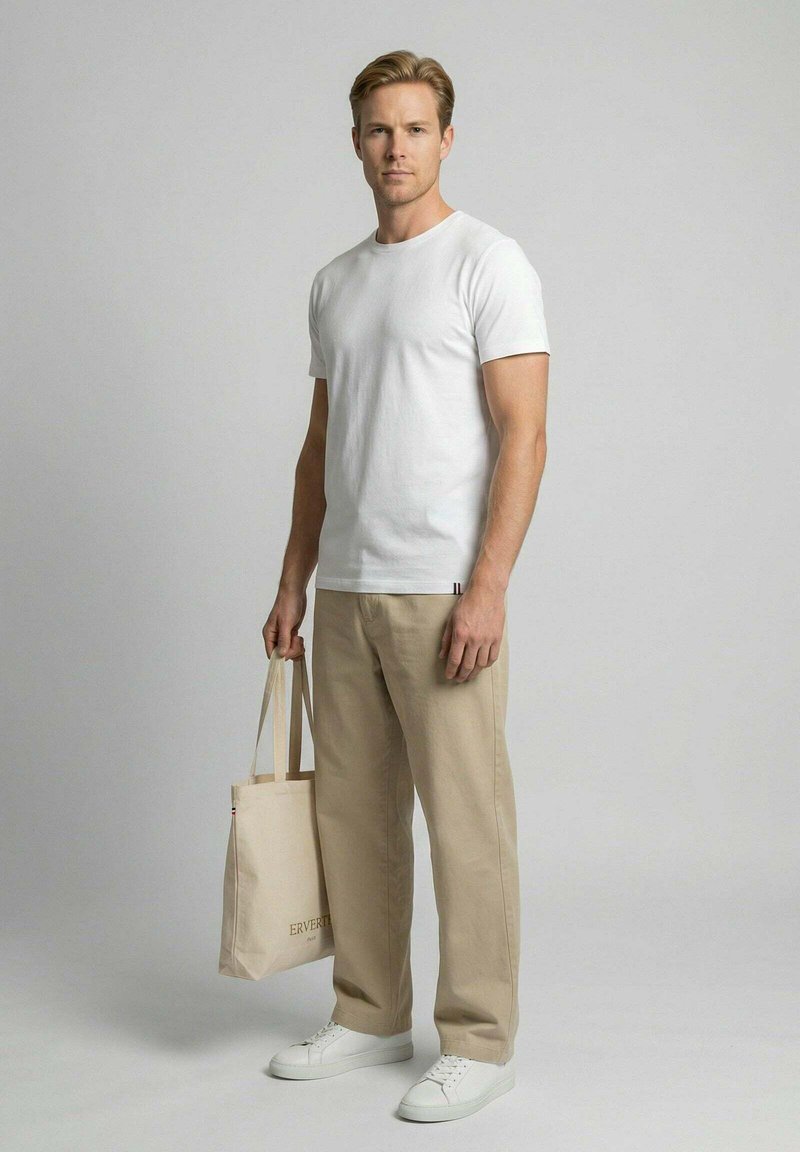 Man wearing white t-shirt, beige pants, white sneakers, holding a beige tote bag, standing against a plain light gray background.