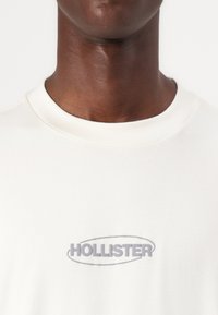 Camiseta de algodón blanca con un logo gris de "HOLLISTER" en el centro. Cuello redondo y textura suave, con detalles cosidos.