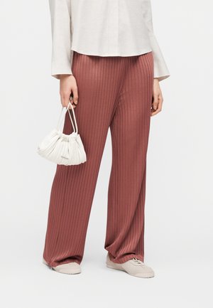 Kvinde iført ribstrikkede, vide bukser i rustfarve, hvid bluse, beige sneakers, holder lille hvid taske med rynker ved siden.