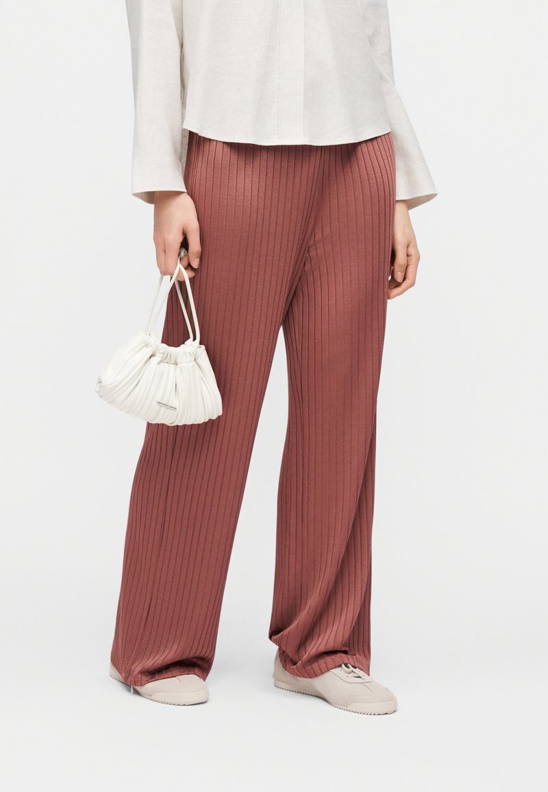 Γυναίκα που φοράει φαρδιά παντελόνια με ανάγλυφο ύφασμα σε σκουριά χρώματος, λευκό πουκάμισο, μπεζ αθλητικά παπούτσια, κρατώντας μικρή λευκή τσάντα με πτυχώσεις στο πλάι.