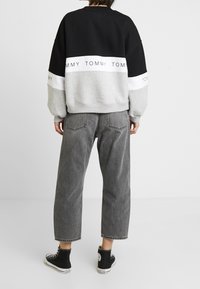 Sudadera a rayas en gris y negro con una banda blanca que presenta texto. Corte holgado, puños acanalados, combinada con unos vaqueros de tiro alto y zapatillas negras.
