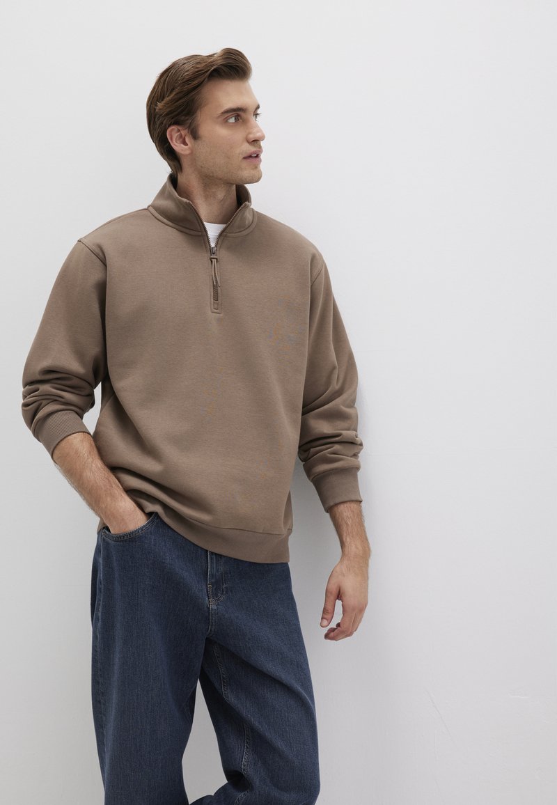 Brown zip-up sweatshirt with ribbed cuffs and hem, paired with loose-fitting blue jeans. The material appears soft and casual.