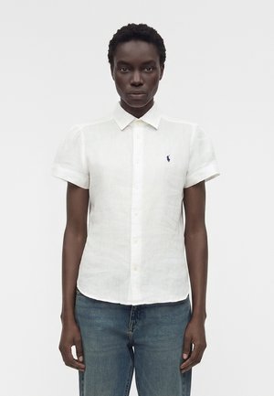 Άτομο που φοράει εφαρμοστή λευκή κοντομάνικη πουκάμισο με μικρό λογότυπο στο στήθος και μπλε τζιν, στέκεται μπροστά σε μονοχρωμο φόντο.