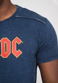 Navy blue t-shirt with a faded texture, featuring a prominent red and yellow "DC" logo on the chest. Short sleeves and a rounded neckline.